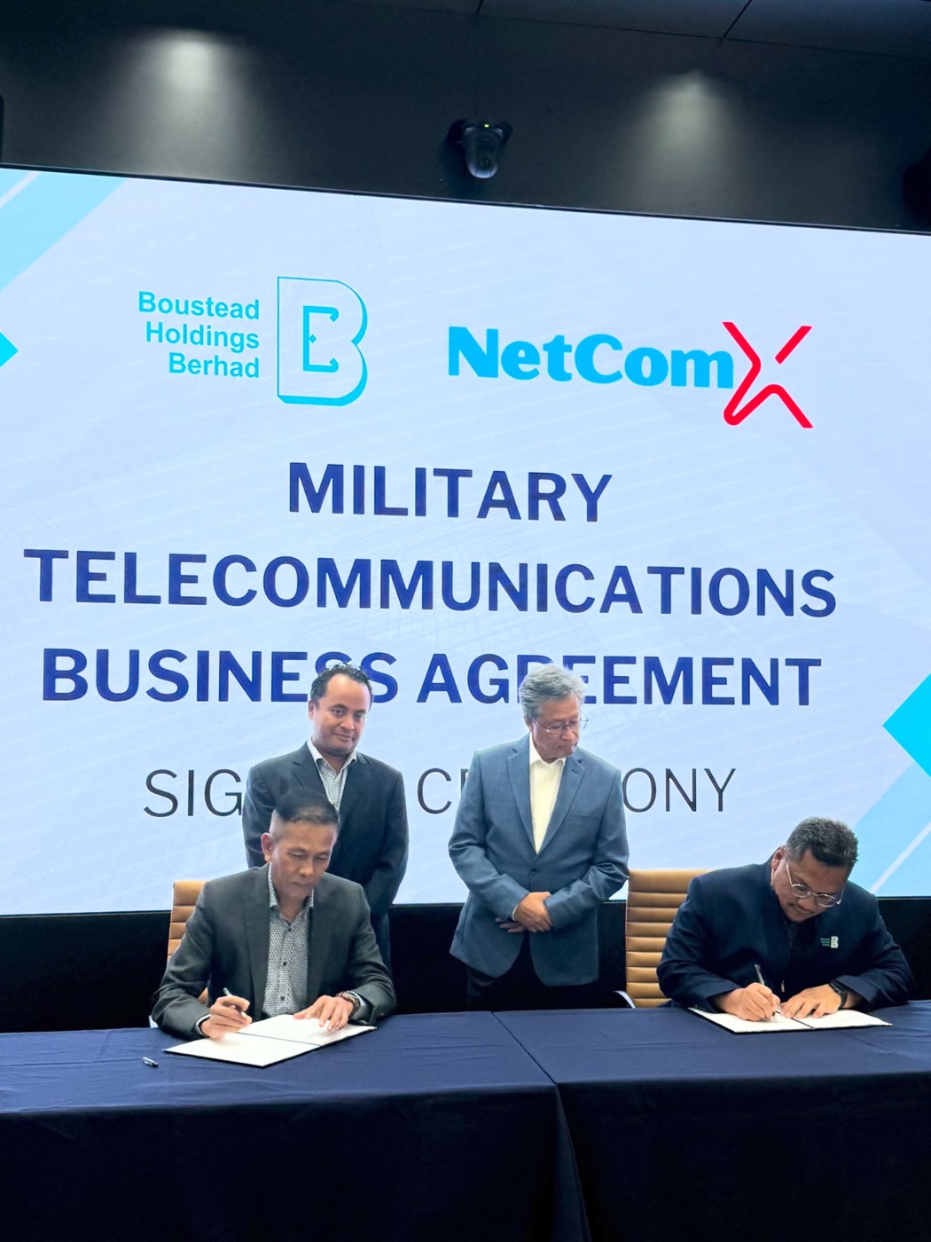 Two seated people signing documents with two people standing behind them.